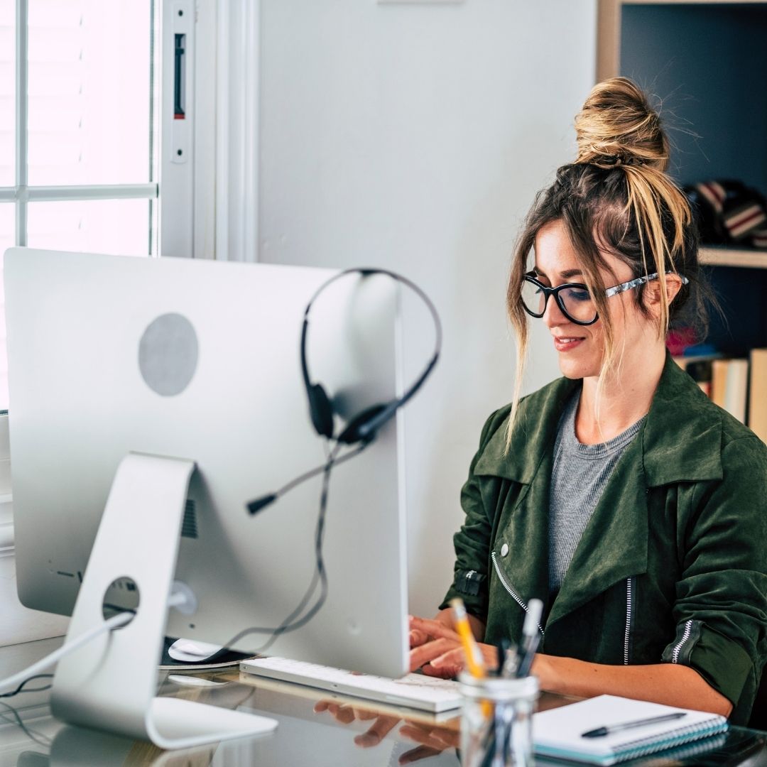 A woman working at a computer with a headset on the corner of the monitor.